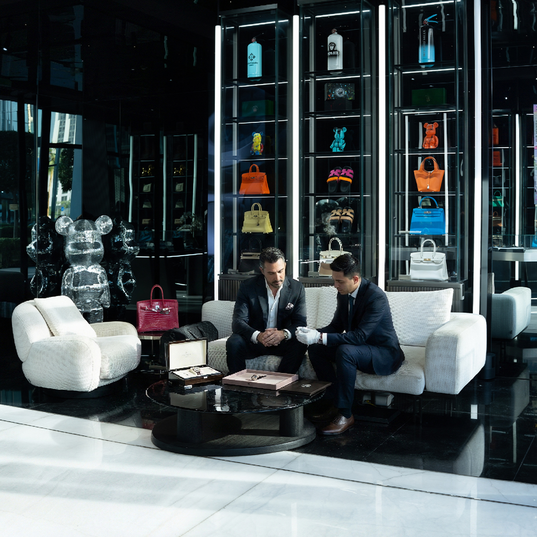 Two men discussing luxury watches in a modern showroom with various designer bags displayed in the background.